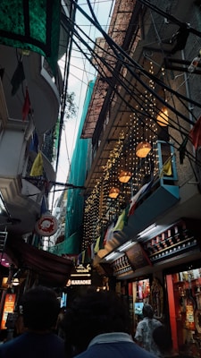 A narrow street crowded with signs, lights, and hanging cables. Colorful flags and decorative lights add vibrancy to the scene, while signage for karaoke and other businesses suggest a lively area.