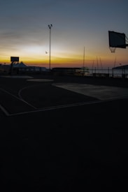 A basketball court is silhouetted against a vibrant sunset. The sky displays a gradient of colors, transitioning from deep blue to bright orange near the horizon. A bird is flying in the distance, adding to the tranquil scene. Streetlights and basketball hoops frame the scene, while the outlines of buildings and poles can be seen near the waterfront.