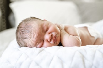 A sleeping newborn baby lies on a soft, white quilt with eyes closed and one arm tucked under the head. The infant's skin appears smooth and delicate, with wisps of fine hair visible on the scalp. Bright, natural light softly illuminates the scene, enhancing the peaceful atmosphere.