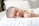 A sleeping newborn baby lies on a soft, white quilt with eyes closed and one arm tucked under the head. The infant's skin appears smooth and delicate, with wisps of fine hair visible on the scalp. Bright, natural light softly illuminates the scene, enhancing the peaceful atmosphere.