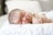 A sleeping newborn baby lies on a soft, white quilt with eyes closed and one arm tucked under the head. The infant's skin appears smooth and delicate, with wisps of fine hair visible on the scalp. Bright, natural light softly illuminates the scene, enhancing the peaceful atmosphere.