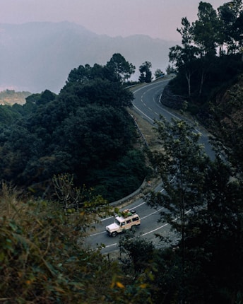 A winding road surrounded by lush greenery and dense forest, with a white SUV carrying gear on its roof driving along it. The background features misty mountains and a cloudy sky, creating an atmospheric scene.