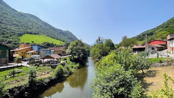A serene landscape featuring a small river flowing through a rural village nestled in a valley. Colorful houses line the banks of the river, surrounded by lush greenery and rolling hills in the background. The sky is clear and blue, allowing sunlight to illuminate the vibrant scene.