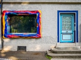 A textured exterior wall featuring a vividly painted, abstract window frame in vibrant red, yellow, and blue hues. Next to the window is a blue door with decorative wrought iron grilles and small steps leading up to it.