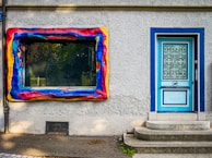 A textured exterior wall featuring a vividly painted, abstract window frame in vibrant red, yellow, and blue hues. Next to the window is a blue door with decorative wrought iron grilles and small steps leading up to it.