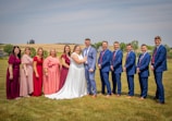 A group of people stands on a grassy lawn. In the center, a couple is dressed in formal wedding attire. Surrounding them are six individuals on the left, dressed in various shades of pink, red, and purple dresses, and five individuals on the right, wearing matching blue suits and red ties. The background features a rural landscape with rolling fields and a few trees.
