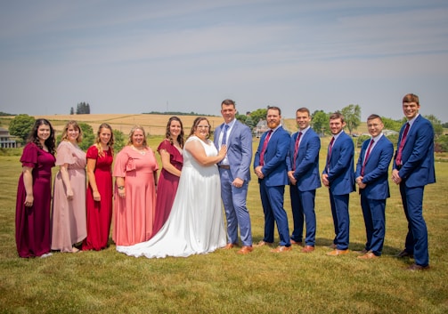 A group of people stands on a grassy lawn. In the center, a couple is dressed in formal wedding attire. Surrounding them are six individuals on the left, dressed in various shades of pink, red, and purple dresses, and five individuals on the right, wearing matching blue suits and red ties. The background features a rural landscape with rolling fields and a few trees.