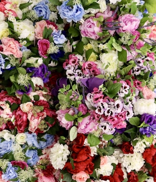 A vibrant array of artificial flowers in various colors and types, including roses, peonies, and hydrangeas. The arrangement includes shades of pink, red, purple, blue, and white, with lush green leaves interspersed throughout.