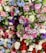 A vibrant array of artificial flowers in various colors and types, including roses, peonies, and hydrangeas. The arrangement includes shades of pink, red, purple, blue, and white, with lush green leaves interspersed throughout.