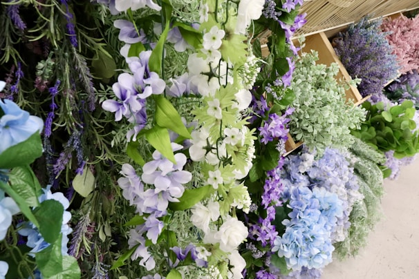 A variety of artificial flowers are arranged closely together, featuring shades of purple, blue, white, and green. The flowers appear to be displayed on shelves or baskets, creating a lush and vibrant floral arrangement.