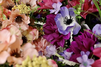 A vibrant assortment of artificial flowers featuring a variety of blooms in shades of pink, purple, and peach against a green leafy background. The petals are delicate and detailed, with some featuring contrasting centers and others accompanied by small buds or ferns.