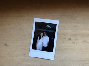 A Polaroid photo rests on a wooden surface, depicting two people standing close together. They both appear to be smiling, wearing white attire, under a structure with beams visible overhead.