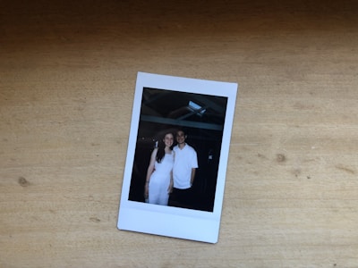 A Polaroid photo rests on a wooden surface, depicting two people standing close together. They both appear to be smiling, wearing white attire, under a structure with beams visible overhead.