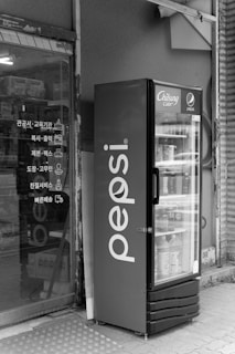 A vending machine with the Pepsi logo is placed outside a store. The machine is filled with various beverages. The store entrance nearby shows signage in Korean, and the glass reflects some products inside. The scene is quiet and appears to be in an urban setting.