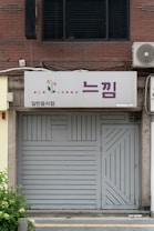 A storefront with a closed gray door and a sign above displaying Korean text. The sign includes a graphic of small colorful flowers and dots, and the surrounding building facade features brickwork. An air conditioning unit is mounted to the right of the sign.