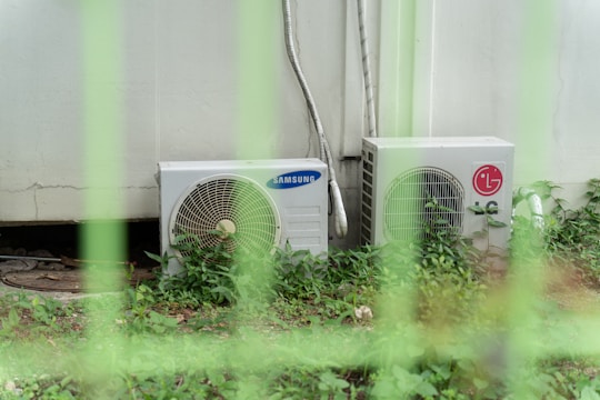 Two outdoor air conditioning units, one branded Samsung and the other LG, are positioned side by side against a light-colored wall. They are surrounded by green plants and have metal pipes extending upward. The scene is partially obscured by green fence bars.