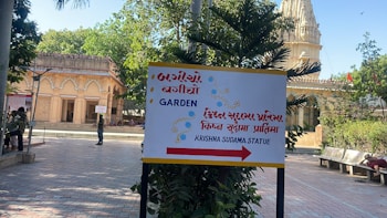 The image depicts an outdoor scene featuring a temple with intricate carvings and a pathway leading towards it. There is a multilingual signboard indicating directions to a garden and the Krishna Sudama statue. People are walking and sitting in the area, with trees and plants providing greenery.
