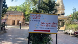 The image depicts an outdoor scene featuring a temple with intricate carvings and a pathway leading towards it. There is a multilingual signboard indicating directions to a garden and the Krishna Sudama statue. People are walking and sitting in the area, with trees and plants providing greenery.