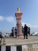 A group of people stands near a tall pedestal featuring a carved pink stone column with a metallic statue on top, resembling the Ashoka Pillar. The setting appears to be a coastal area with parked cars and a clear blue sky in the background.