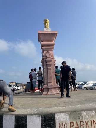 A group of people stands near a tall pedestal featuring a carved pink stone column with a metallic statue on top, resembling the Ashoka Pillar. The setting appears to be a coastal area with parked cars and a clear blue sky in the background.