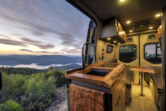 A cozy and modern camper van interior with wooden countertops and a sleek design. The view from the van's window overlooks a scenic landscape with a lake surrounded by hills and forests under a partly cloudy sky at sunset.