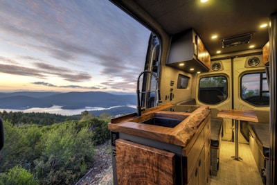 A cozy and modern camper van interior with wooden countertops and a sleek design. The view from the van's window overlooks a scenic landscape with a lake surrounded by hills and forests under a partly cloudy sky at sunset.