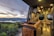 A cozy and modern camper van interior with wooden countertops and a sleek design. The view from the van's window overlooks a scenic landscape with a lake surrounded by hills and forests under a partly cloudy sky at sunset.