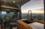 Interior view of a camper van featuring modern design elements, including a driver's seat and dashboard with a built-in display. The foreground shows a wooden countertop with a sleek black faucet. Through the large window, a scenic landscape with rolling hills and a setting sun is visible, providing a serene, natural backdrop.