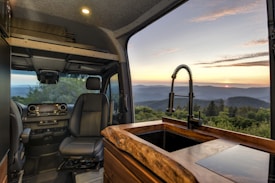 Interior view of a camper van featuring modern design elements, including a driver's seat and dashboard with a built-in display. The foreground shows a wooden countertop with a sleek black faucet. Through the large window, a scenic landscape with rolling hills and a setting sun is visible, providing a serene, natural backdrop.