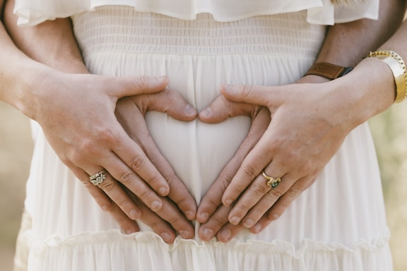 Four hands gently rest on a pregnant belly, forming a heart shape. The hands belong to two individuals, each wearing rings. The clothing is light and airy, suggesting a sense of warmth and tenderness.