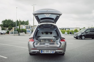 A silver car is parked in an open parking lot with its trunk open, revealing a complex interior setup possibly for equipment or cargo organization. The surrounding area includes several buildings, trees, and another black vehicle is parked nearby.