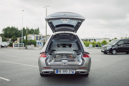 A silver car is parked in an open parking lot with its trunk open, revealing a complex interior setup possibly for equipment or cargo organization. The surrounding area includes several buildings, trees, and another black vehicle is parked nearby.
