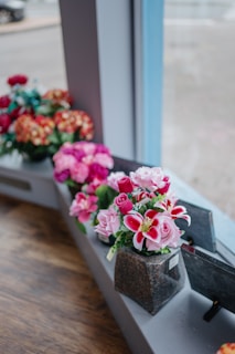 A row of artificial flower arrangements in various colors displayed on a windowsill. The flowers include pink roses and vibrant red lilies, crafted with detailed petals and green leaves. Each arrangement is neatly placed in a small square vase made to look like stone, positioned along a wooden surface. The background shows a blurred outdoor scene through the window glass.