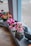 A row of artificial flower arrangements in various colors displayed on a windowsill. The flowers include pink roses and vibrant red lilies, crafted with detailed petals and green leaves. Each arrangement is neatly placed in a small square vase made to look like stone, positioned along a wooden surface. The background shows a blurred outdoor scene through the window glass.