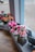 A row of artificial flower arrangements in various colors displayed on a windowsill. The flowers include pink roses and vibrant red lilies, crafted with detailed petals and green leaves. Each arrangement is neatly placed in a small square vase made to look like stone, positioned along a wooden surface. The background shows a blurred outdoor scene through the window glass.