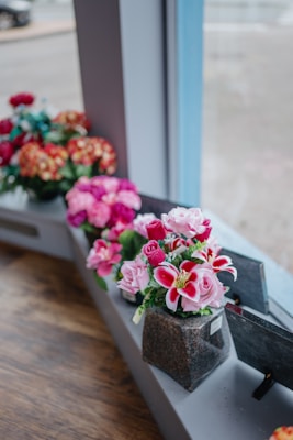 A row of artificial flower arrangements in various colors displayed on a windowsill. The flowers include pink roses and vibrant red lilies, crafted with detailed petals and green leaves. Each arrangement is neatly placed in a small square vase made to look like stone, positioned along a wooden surface. The background shows a blurred outdoor scene through the window glass.