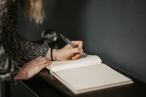 An individual is writing in a notebook with a pen. The person is wearing a dark patterned long-sleeve shirt and has jewelry on. The scene has a soft, introspective feel with a blurred background and gentle lighting.