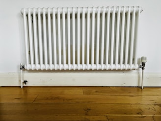 A white, wall-mounted radiator with vertical bars is fixed to a plain white wall above a light wooden floor. The baseboard and pipe fittings are also visible, with some signs of wear and discoloration on the radiator.