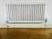 A white, wall-mounted radiator with vertical bars is fixed to a plain white wall above a light wooden floor. The baseboard and pipe fittings are also visible, with some signs of wear and discoloration on the radiator.
