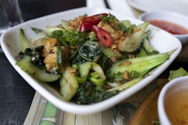 A dish featuring sautéed green vegetables, likely bok choy, mixed with sliced red chili peppers, scallions, and pieces of tofu. The ingredients are presented in a white, rectangular-shaped plate and are garnished with crispy fried onions. A small bowl with a red dipping sauce is visible in the background, adding a hint of spice to the presentation.