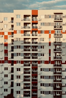 A multi-story residential building with a symmetrical facade featuring rectangular balconies and numerous windows. The structure is painted in a combination of beige, red, and gray, with air conditioning units visible on most balconies. The sky is visible in the background with a few clouds.
