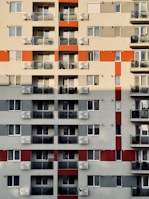 A high-rise residential building with multiple floors featuring a grid pattern. Each floor has several units with glass balconies and assorted chairs visible. The facade includes alternating red and grey sections, adding a geometric visual interest.