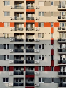 A high-rise residential building with multiple floors featuring a grid pattern. Each floor has several units with glass balconies and assorted chairs visible. The facade includes alternating red and grey sections, adding a geometric visual interest.