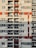 A high-rise residential building with multiple floors featuring a grid pattern. Each floor has several units with glass balconies and assorted chairs visible. The facade includes alternating red and grey sections, adding a geometric visual interest.