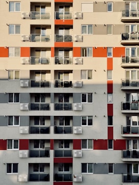 A high-rise residential building with multiple floors featuring a grid pattern. Each floor has several units with glass balconies and assorted chairs visible. The facade includes alternating red and grey sections, adding a geometric visual interest.