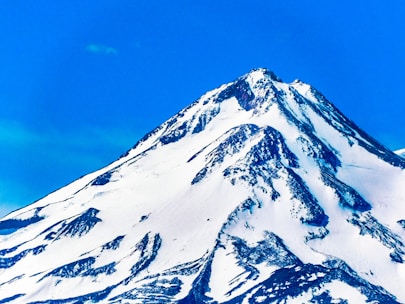 A majestic snow-covered mountain peak rises sharply against a vibrant blue sky. The mountain features rugged terrain with patches of black rock visible through the white snow, creating stark contrasts.