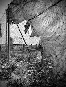 A black and white image of a damaged chain-link fence with torn sections. Behind the fence, there are signs of neglect with scattered debris and overgrown plants. In the background, graffiti can be seen on a wall, indicating an urban setting. The perspective creates a feeling of looking through confinement or barriers.