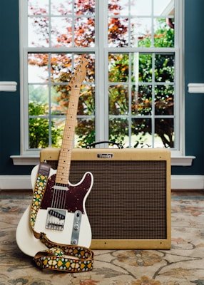 A white electric guitar with a colorful strap leans against a vintage-style Fender amplifier. The setup is placed on a patterned rug in a room with large windows, through which greenery and autumn-colored trees are visible.