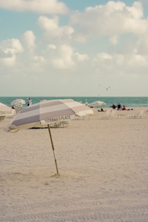 A sandy beach setting with a large umbrella labeled 'Esteban's Place' positioned prominently in the foreground. Several people are seen relaxing on the beach in the background near the shoreline. The sky is partly cloudy, and the ocean water is visible in the distance.