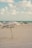 A sandy beach setting with a large umbrella labeled 'Esteban's Place' positioned prominently in the foreground. Several people are seen relaxing on the beach in the background near the shoreline. The sky is partly cloudy, and the ocean water is visible in the distance.